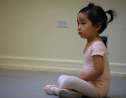 A young dancer in pink sits cross-legged with hands in her lap.