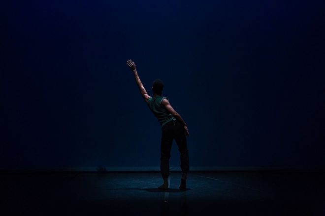 A dancer reaches his arm on a dark stage